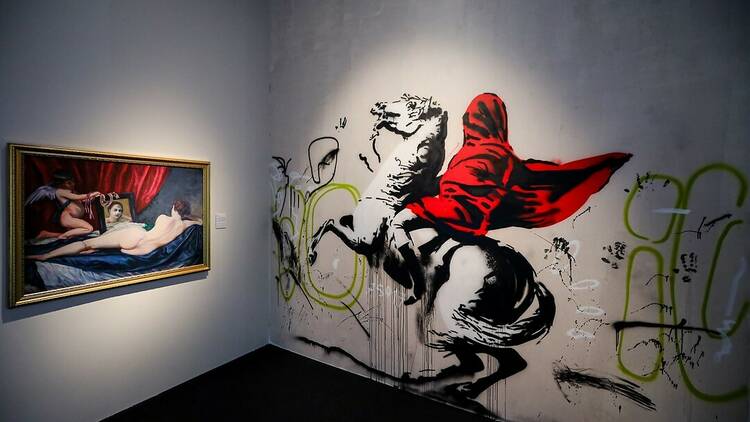 A Banksy mural featuring a red-cloaked figure on a horse in a gallery space.