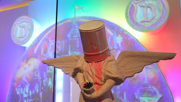 A sculpture of an angel with a bucket on its head in front of a rainbow projection.