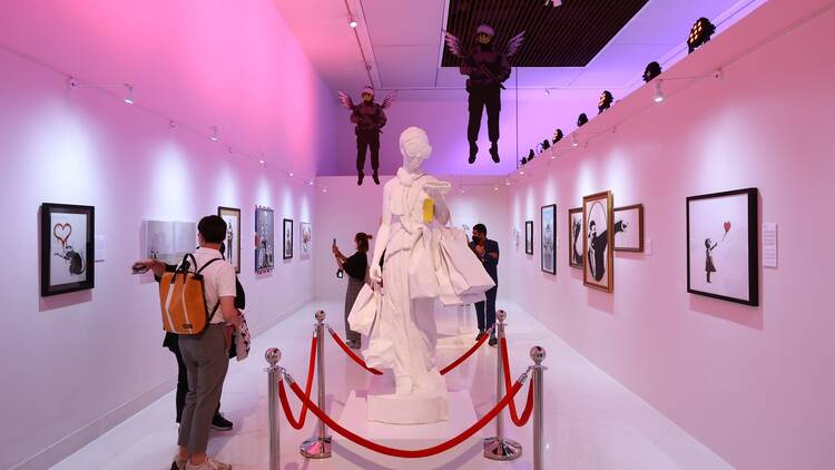 A white sculpture in a purple-lit gallery space filled with Banksy artworks.