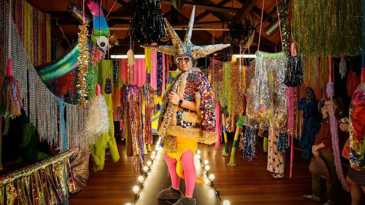 A person in an outlandish rainbow outfit with a gold headdress stands in a colourful room.