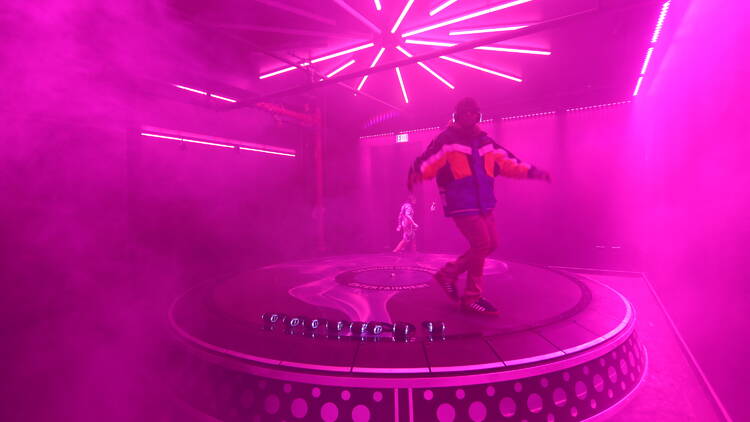 A person dances on an oversized record player, cast in pink light and haze.