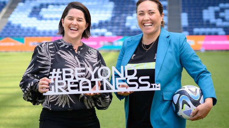 Two women standing in a stadium holding a soccer ball and a sign that says: #BEYOND GREATNESS