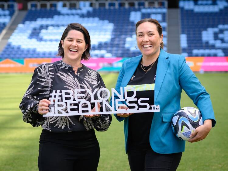 Two women standing in a stadium holding a soccer ball and a sign that says: #BEYOND GREATNESS