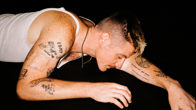 A person with a moustache and tattoos and wearing a white singlet top lays peacefully on a dark ground