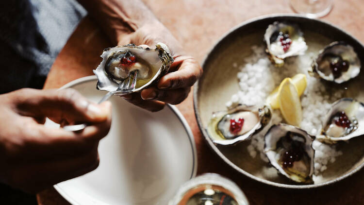 A person using a small fork to get an oyster, while a plate of oysters sits on a table in the background.