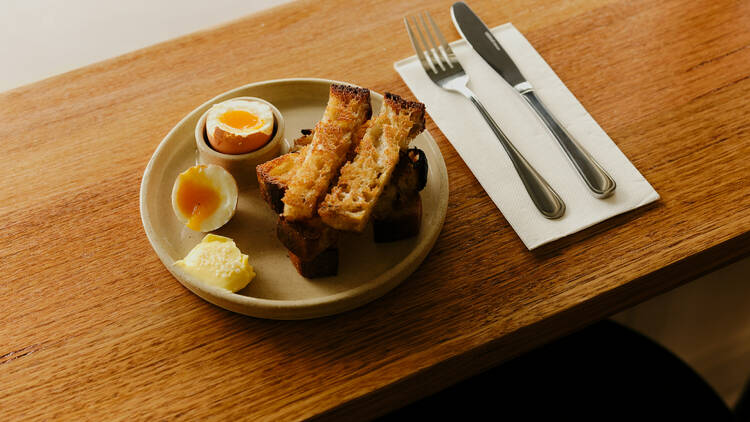 A plate of boiled eggs and soldier toast with cutlery on a wooden bench.
