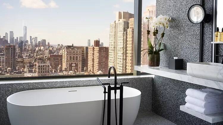 A soaking tub in a modern bathroom rests just under a large window with a view of the Manhattan skyline