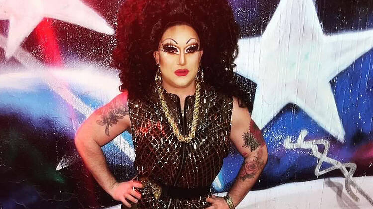 Alotta McGriddles, a drag performer, wearing a curly brunette wig and a gold-and-black romper.