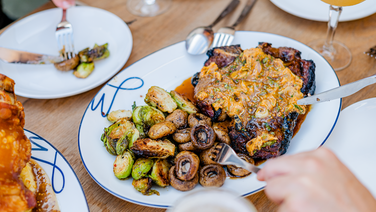 Hands using knives and forks to serve up a big steak covered in sauce, served with mushrooms and Brussels sprouts.