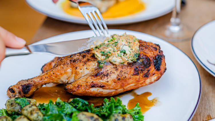 Hands using a knife and fork to serve up grilled chicken topped with buttery sauce, served with gnocchi and kale.