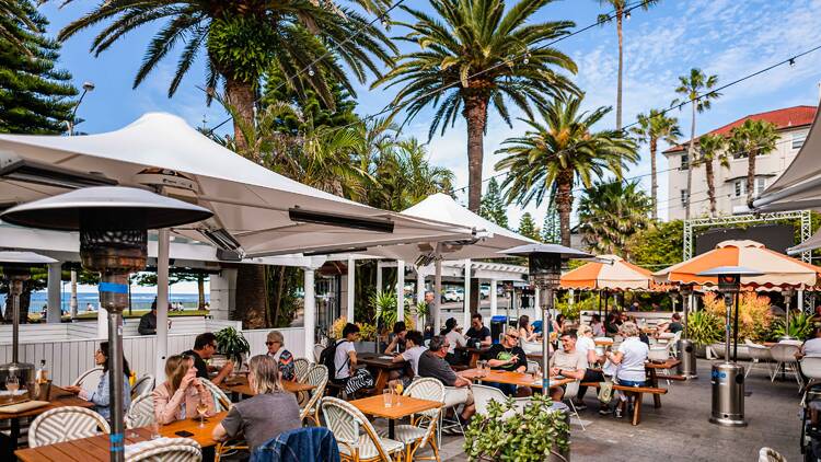 People sitting in a beer garden, under umbrellas and palm trees.