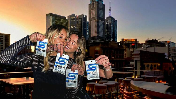 Two women stand close together holding packets of Zuum on the rooftop of Her bar, with the Melbourne skyline in the background.
