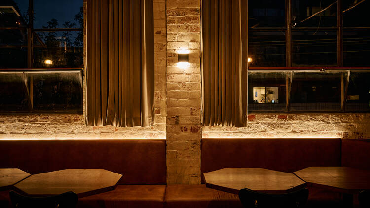 The view out of curtain-framed windows from within a dimly lit bar of hexagonal tables, exposed brick walls and leather seating.   