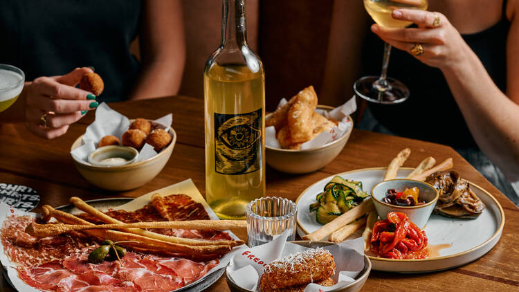 A table of food including a plate of charcuterie, breadsticks and olives; a bottle of white wine; assorted antipasti, arancini and a small ramekin of white sauce; and the arms and hands of a pair of guests in the background.