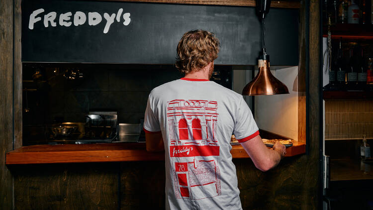 The back of a waiter in a red graphic T-shirt taking a plate of food from the kitchen countertop in a dimly lit bar