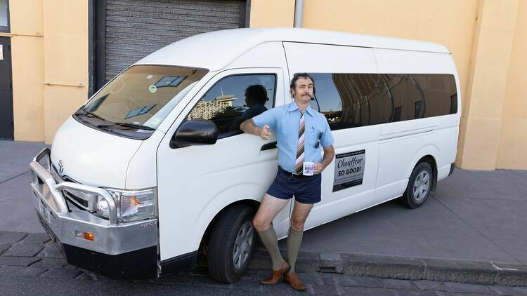 A man in short shorts, a tie and a headset poses with his thumbs up in front of a white mini van.