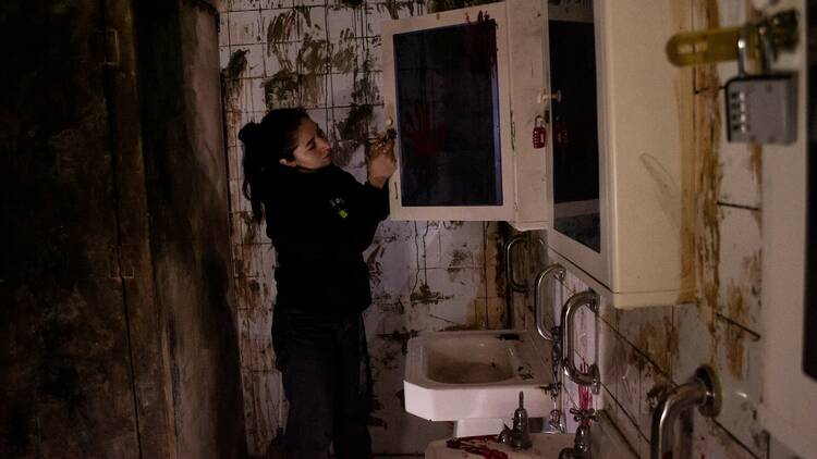 A person tinkers with a lock on a cupboard with in a bathroom covered in dirt and blood.