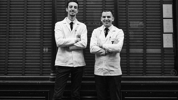 Two bartenders in white coats and black ties standing next to one another with crossed arms and smiling at the camera.
