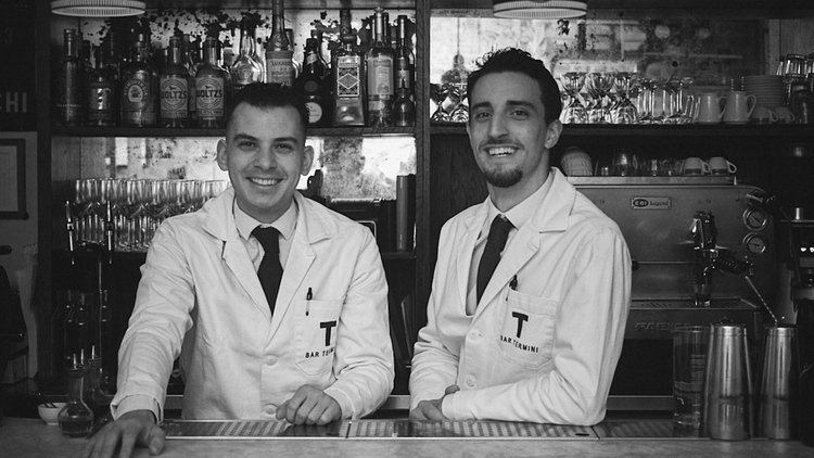 Two bartenders in white coats and black ties standing next to one another behind the bar and smiling at the camera.
