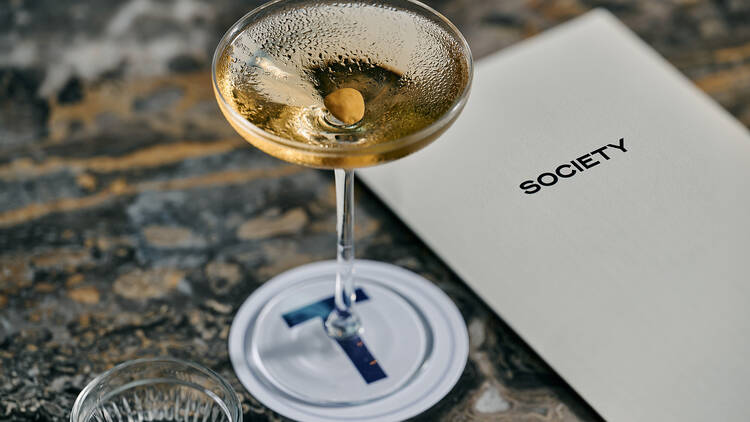 Cocktail and glass of water sitting next to a 'Society' menu on a bar top.