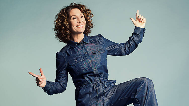 The comedian Kitty Flanagan poses with her fingers in the air while wearing a denim jumpsuit.