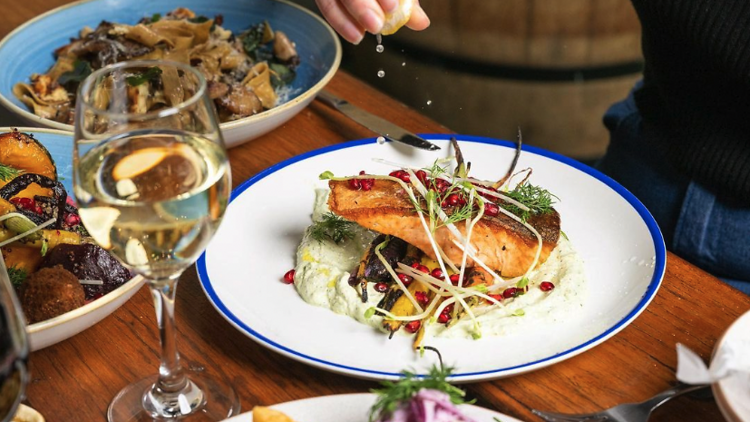 Hand squeezing lemon quarter onto a plate of seared salmon and vegetables with a glass of white wine and other plates of food on a brown wooden table.