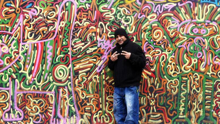 Artist Angel Ortiz poses by a wall of colorful artwork.