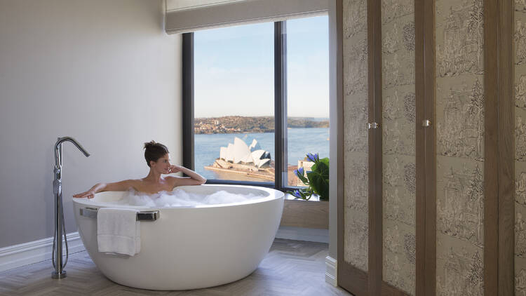 A woman in a bath looking out a window that looks out onto the Sydney Opera House.