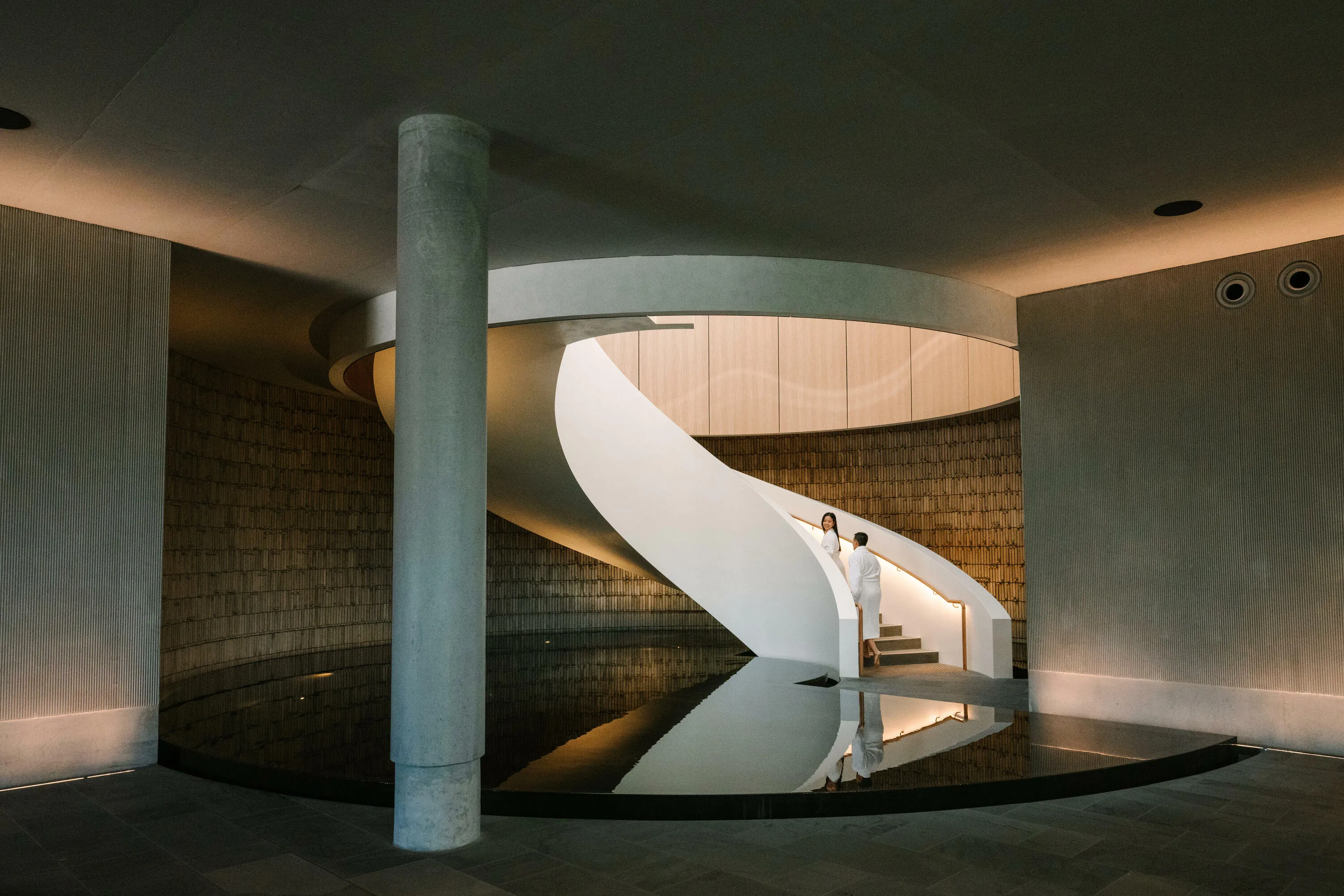Two people in white bath robes walk up a spiral staircase with a water base.