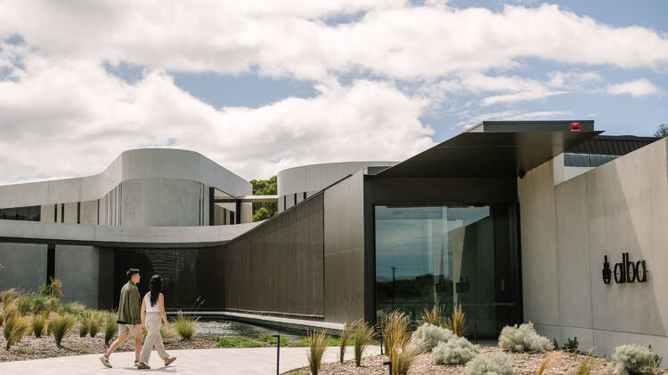 Two people walk into a modern, concrete building surrounded by shrubbery.