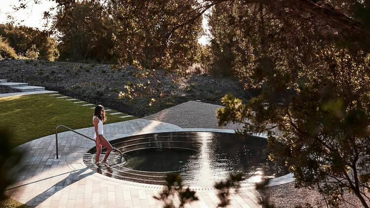 A woman in a white bathing suit walks into a circular-shaped outdoor thermal pool.