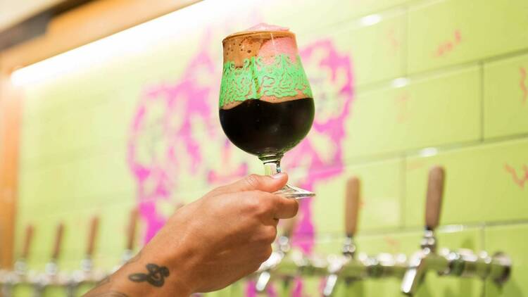 A hand lifting a glass of beer with a green wall with hot pink artwork in the background. 