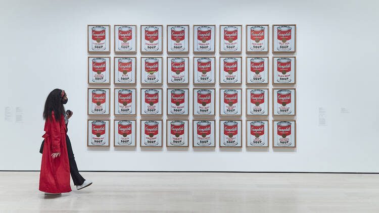 A woman in a red coat walks by Warhol's soup can artwork.