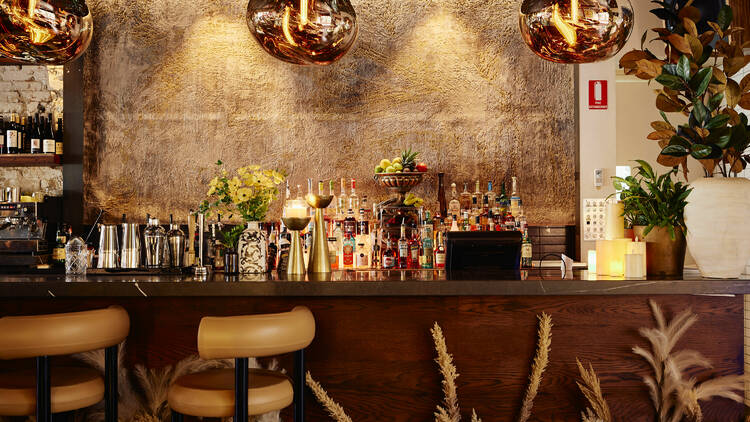 Glamorous, warmly lit wine bar with bottles of liquor, plants and two bar seats. 