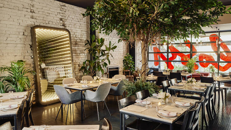 Decorated dining space housing tables, chairs, large plants, and a mirror framed with lights.