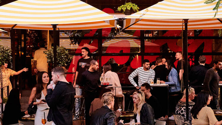 Outdoor patio dining space with orange and white striped umbrellas, lights and revellers seated at tables and standing.