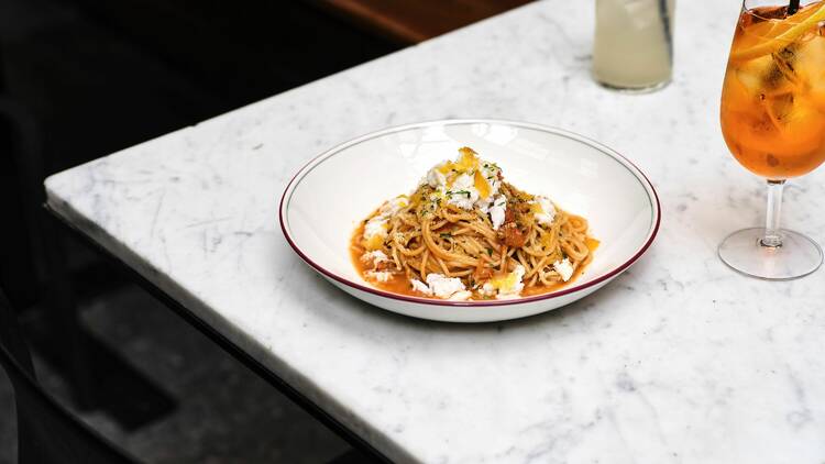 A bowl of spanner crab spaghettini next to an Aperol spritz and another cocktail on a white marble table.