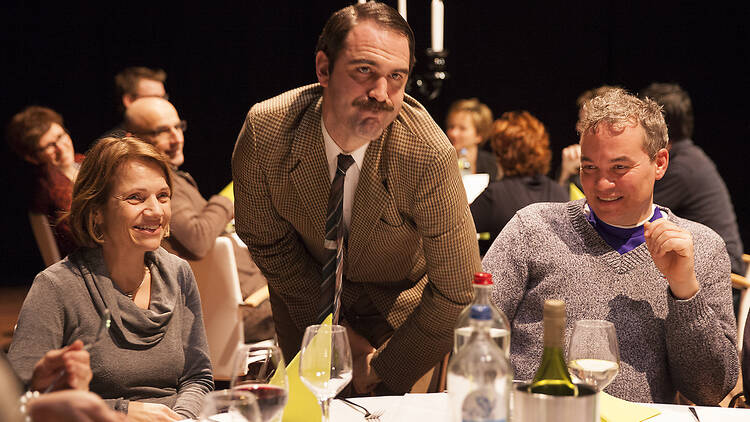 An actor who looks like Basil Faulty pulling a face in front of a table of diners.