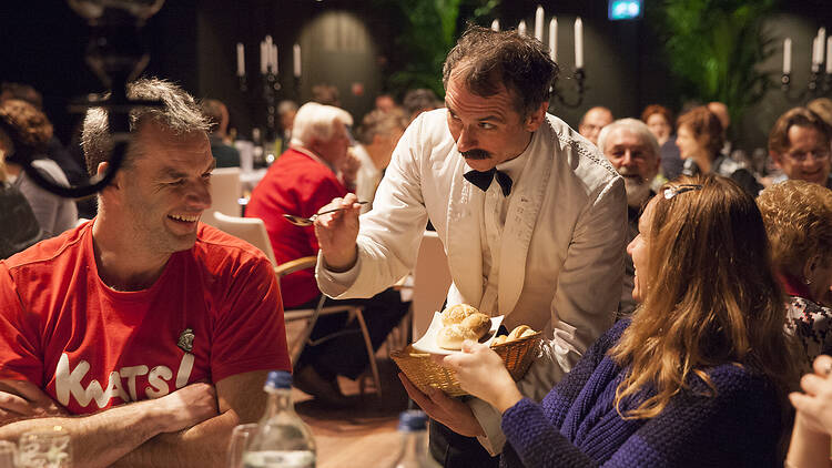 An actor who looks like Manuel from Faulty Towers trying to feed a diner with a spoon.