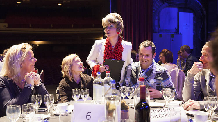 An actor who looks like Sybil Fawlty talking to a table full of diners.