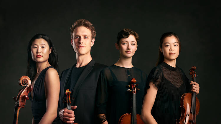 Four musicians all in black hold their instruments while standing against a black backdrop.
