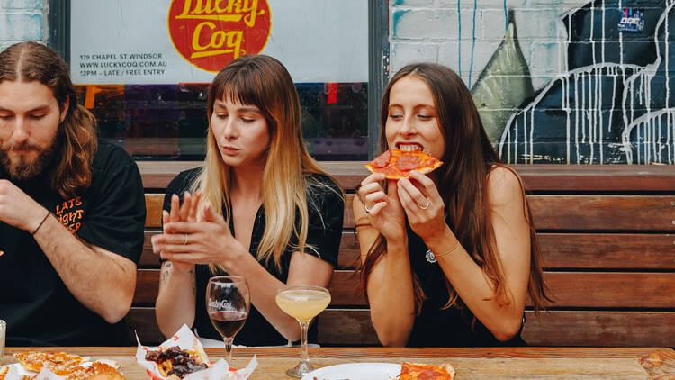 Two women and a man enjoying pizza and drinks at a wooden table.