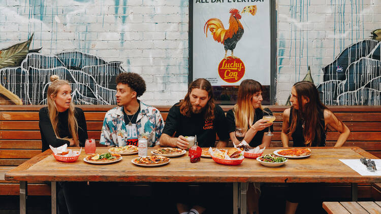 Three women and a man sitting at a table laid out with pizza and drinks in the foreground of a wall covered in graffiti art.