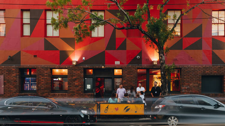 Red-lit building with geometric triangle shapes in the background of a tree and a fast-moving car.