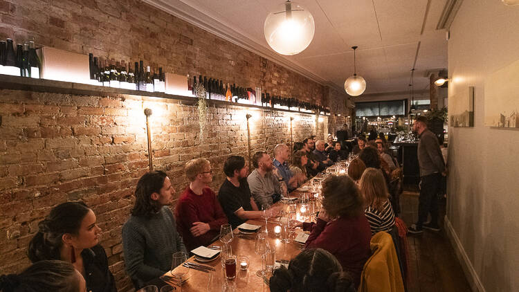 Table of people gathering around a dining table in a narrow room with dim lighting.