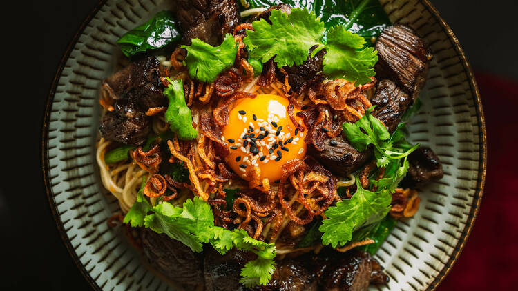 A bowl of noodles topped with cubes of beef, fresh green herbs and a raw egg yolk.