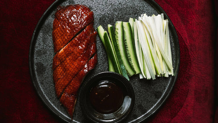 Roast duck sliced on a plate with cucumber sticks and a side of sauce.