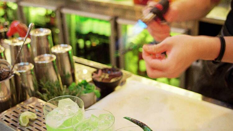 Bartender blowtorching three lime green cocktails at the bar.