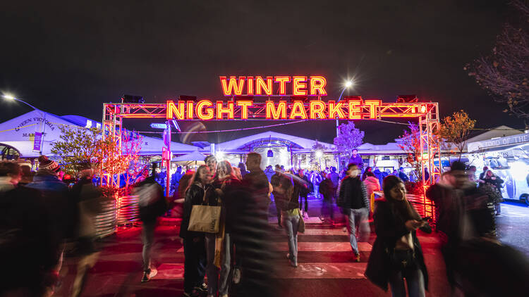 A neon red sign that says 'Winter Night Market' with people walking around.