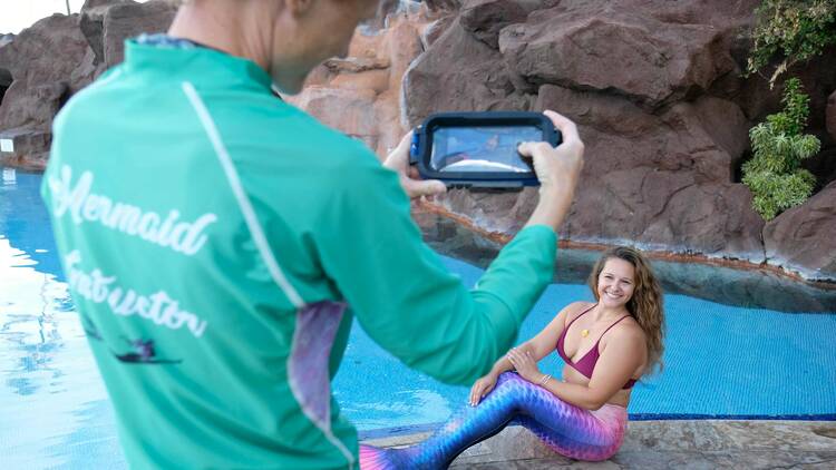 A mermaid poses on the edge of the pool with a smile as someone takes her photo